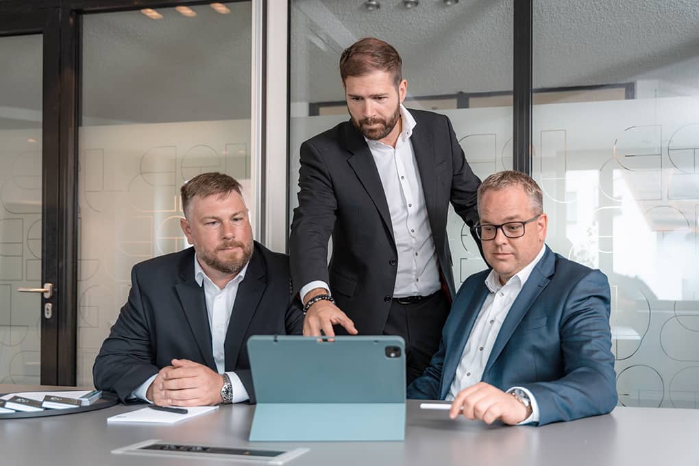 Die drei Geschäftsführer von J2B schauen auf ein Tablet, das vor ihnen auf einem Tisch steht